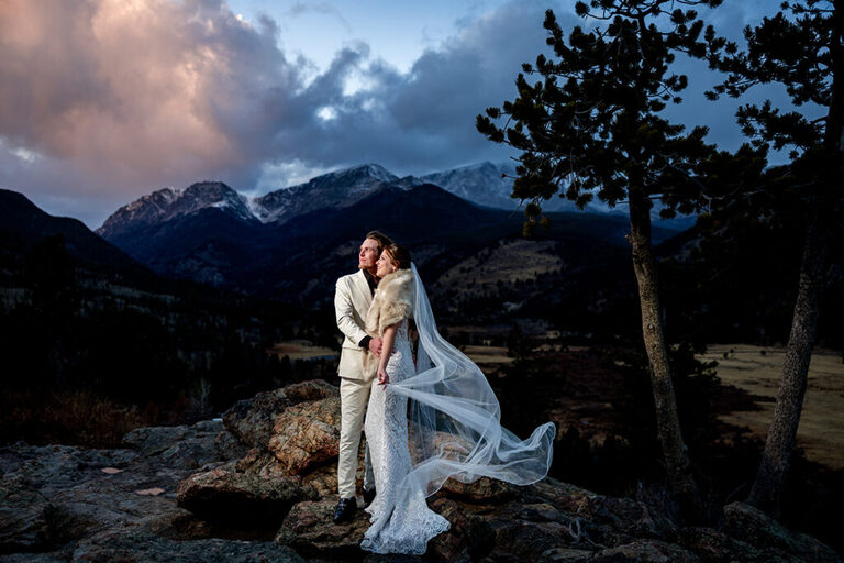a bride and groom at sunset on a rocky outcropping for wedding photos.  The sun is setting behind large mountains behind them.